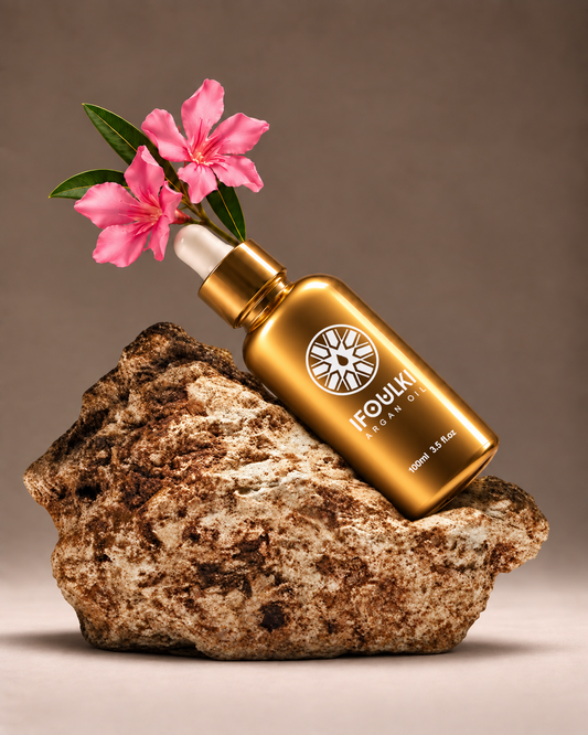 Ifoulki Argan Oil 100ml gold bottle resting on a natural rock beside pink flowers